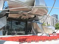 A large bandshell stage, closed off by a glass door, is framed by large curved plates of shiny metal. A large number of red seats are in front of the stage, with a metal trellis forming triangular and diamond shapes above. Large buildings are in the background at right.