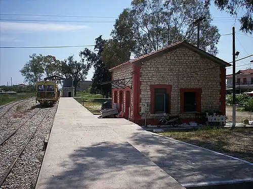 A train approaches a rail station consisting of a small brick building, with a concrete platform between it and the rail road tracks