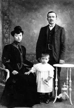 A woman sitting in a long black dress, next to a young child holding a bugle and wearing a white gown, with a man standing up and wearing a suit