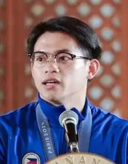 An image of Carlos Yulo speaking to a microphone, wearing his Olympic uniform.
