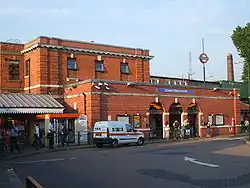 A red-bricked building with a blue sign reading "GOLDERS GREEN STATION" in white letters and people in front all under a blue sky with white clouds