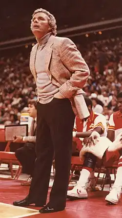 A man with curly gray hair, wearing a tan jacket and shirt with black pants and shoes, standing with his hands in his pockets