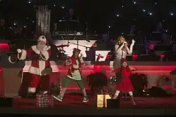 A woman wearing a long white dress. She has long golden hair and is holding a sparkling microphone. She is standing on a large red stage, surrounded by dancers in Santa Claus outfits and generally festive attire.