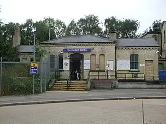 A beige-bricked building with a rectangular, dark blue sign reading "MILL HILL STATION" in white letters all under a blue sky with white clouds
