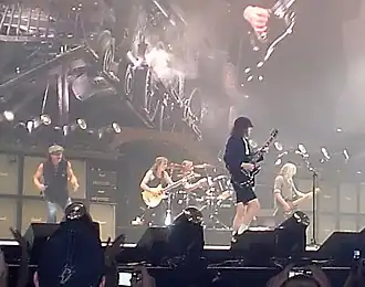 Rock band in performance on a well-lit but hazy stage. we see two guitarists, a bassist, a vocalist off to one side, and a drummer in the rear.