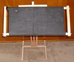 مهره‌دوزی در حین ساخت: سنجاق سینه‌ای از مروارید و کریستال سربی روی پارچۀ اولتراسوید که به قاب گل‌دوزی طوماری متصل شده است