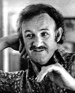 Black-and-white portrait of a middle-aged white man with dark hair and moustache, wearing a patterned collar shirt, resting his hand on his head and looking over to the side.