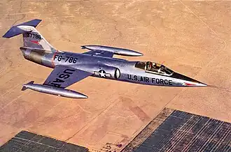 Jet fighter in metallic scheme with T-tail and short wings flying above desert and black constructions