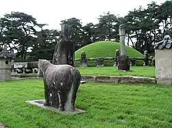 A stone sculpture of a horse and a man, a stone column and a grass covered mound surrounded by a carved stone fence.