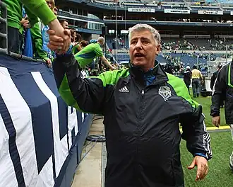 A man in a green and black tracksuit shaking hands with fans at a stadium.
