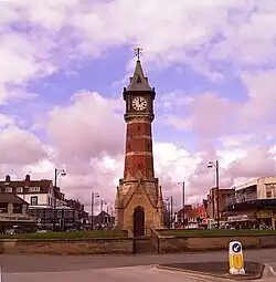 Octagonal brick and limestone tower on a square limestone base, designed like a gothic church porch. The tower is capped with a square lantern, a white clock face on each side, heavy with Roman numerals. The towering roof is capped with a gold-coloured weathervane. The viewpoint is on the short coast road called 'Tower Esplanade', behind the tower is the row of tourist-trap shops in Lumley road. The tower dominates the picture, standing out of a partly blue sky mostly filled with white, purplish, clouds. There is a ring of brick flowerbeds ringing the roundabout the tower is in. The photo was taken in May, the flower beds are just green.